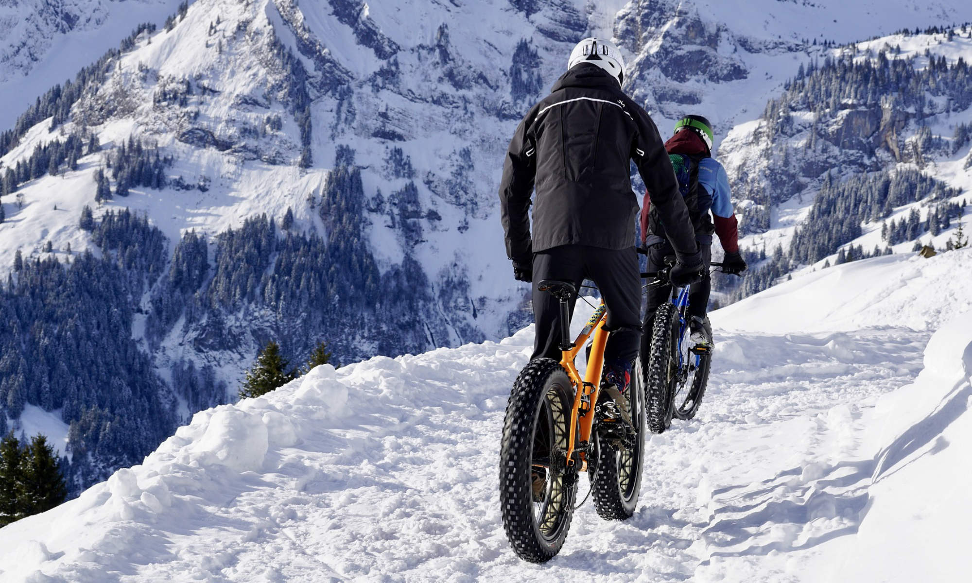 2 Radfahrer mit Fatbikes fahren durch den Schnee, im Hintergrund verschneite Berge.