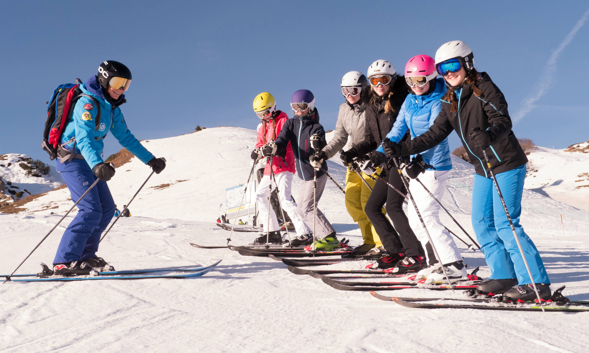 Eine Skilehrerin beim Skikurs mit Teenagern auf Skipiste bei sonnigem Wetter.
