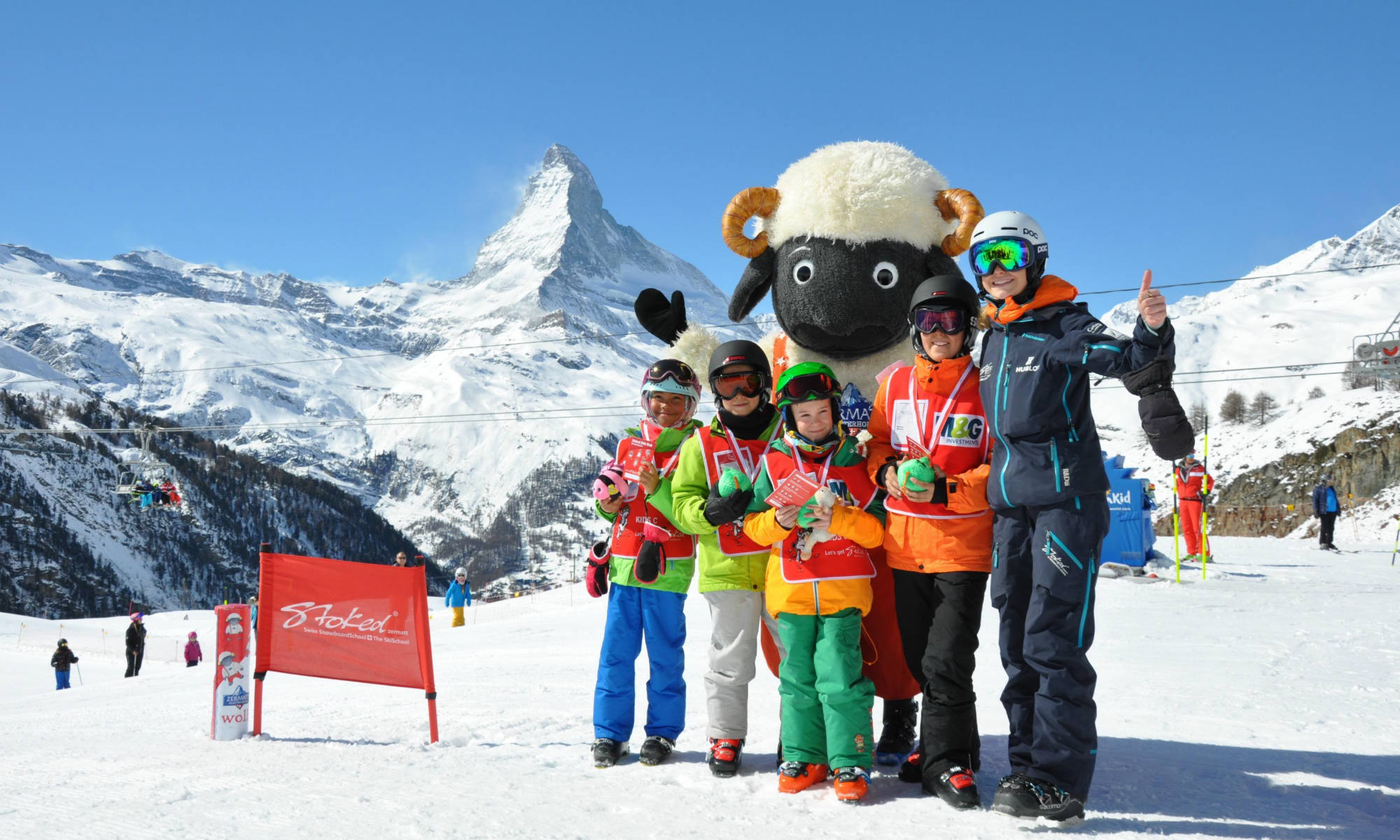 Eine Gruppe Kinder und eine Skilehrerin stehen auf einer Skipiste vor dem Panorama des Matterhorns.