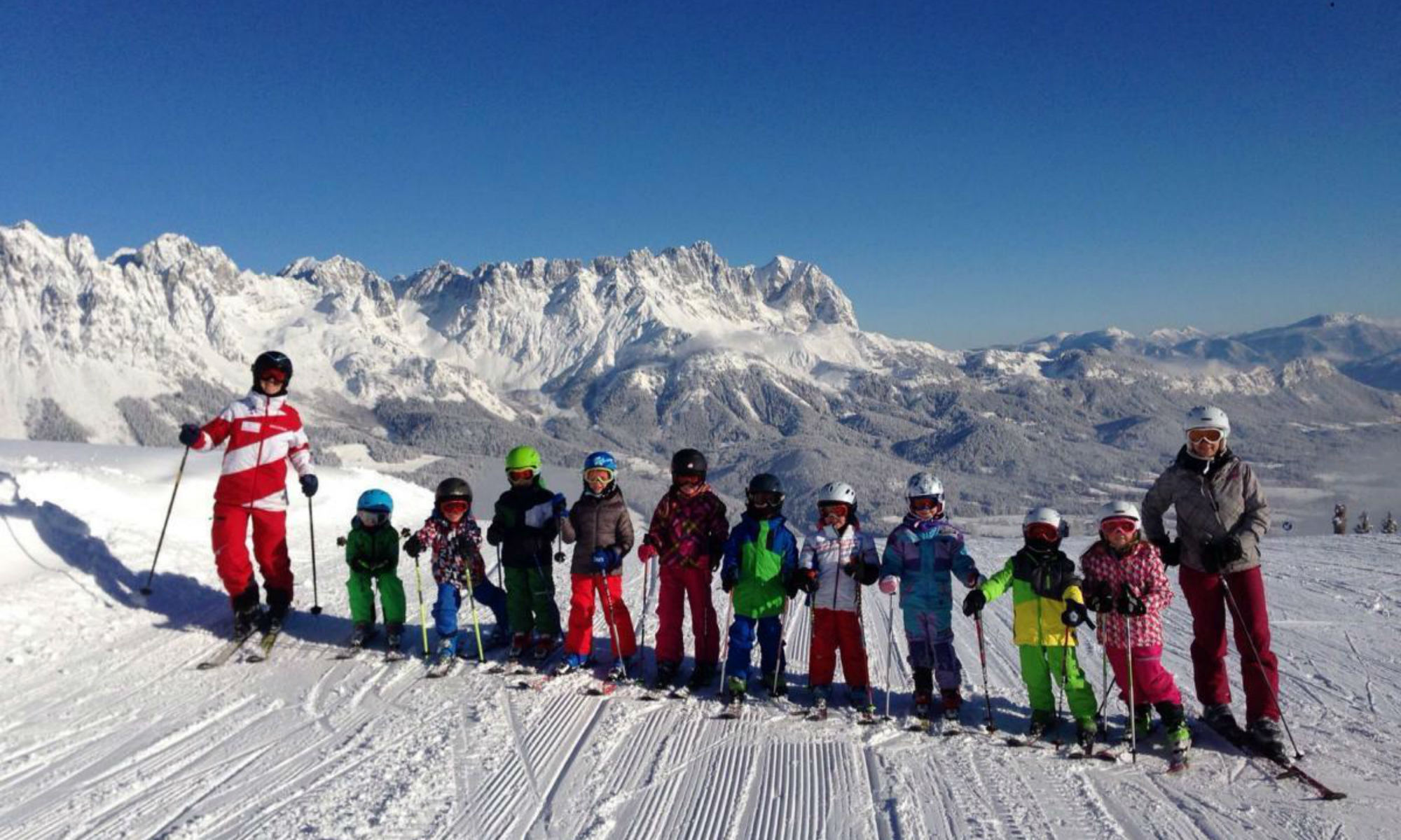 Kindergruppe mit Skilehrer beim Kurs auf der Piste mit Bergpanorama.