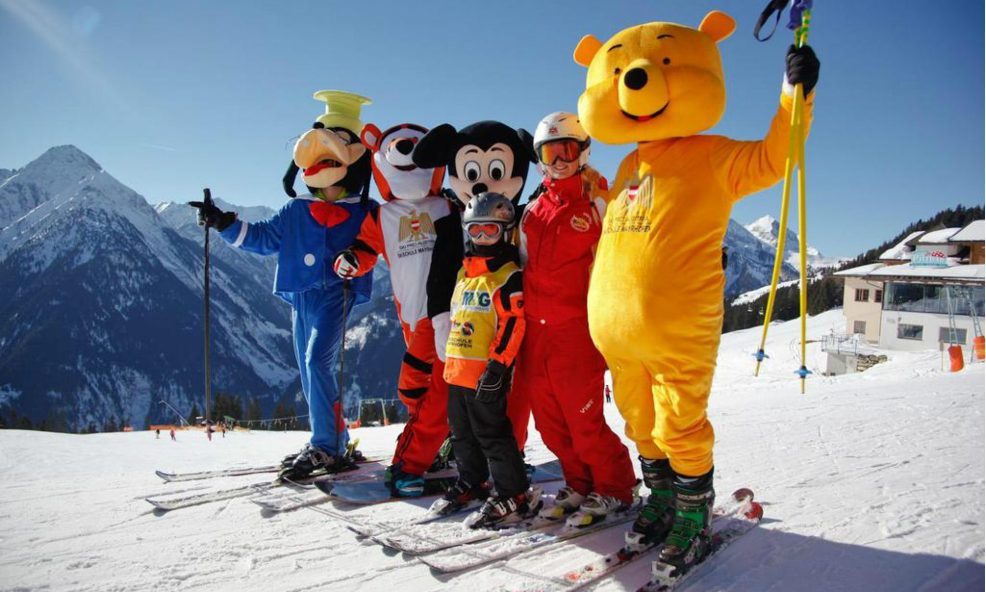 4 Maskottchen der Skischule Mayrhofen Ski Pro Austria stehen mit einer Skilehrerin und einem Kind auf einer sonnigen Piste im Skigebiet Mayrhofen.