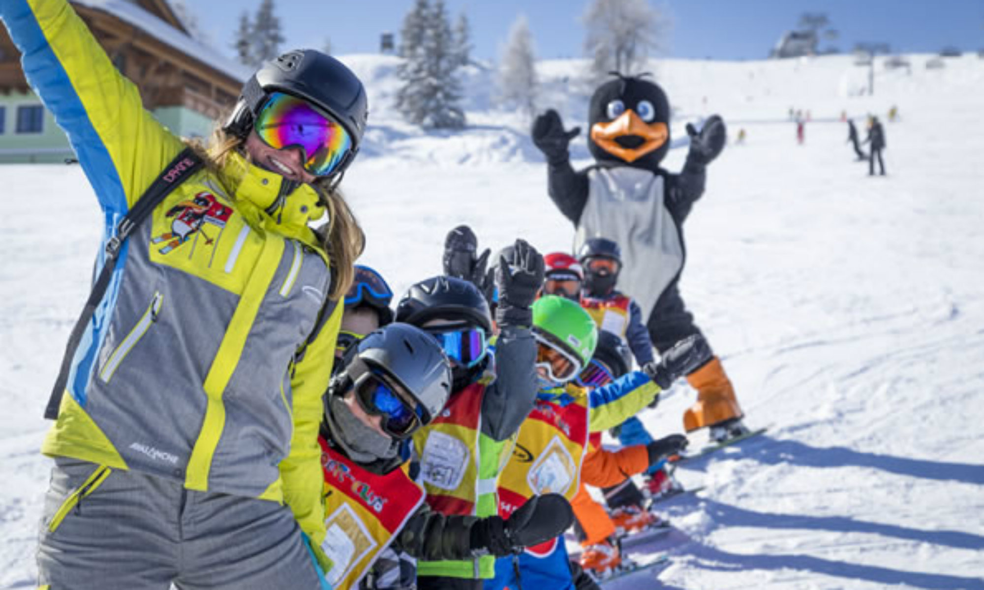 Eine Skilehrerin der Skischule Sport am Jet Flachau, eine Gruppe Kinder und ein Pinguin Maskottchen stehen auf einer sonnigen Piste im Skigebiet Flachau.