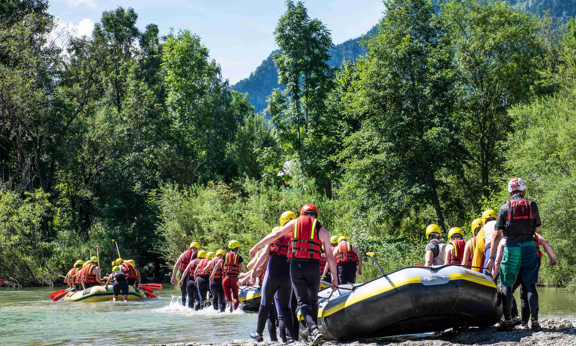 Mehrere Gruppen tragen Schlauchboote zur Einstiegsstelle am Fluss.
