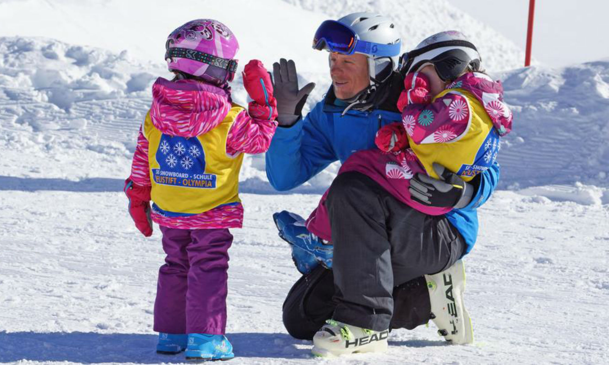 Ein Skilehrer klatscht mit einem Kind auf einer sonnigen Piste am Stubaier Gletscher ab, während er ein zweites Kind im Arm hält.