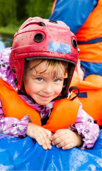 Ein kleines Mädchen sitzt mit Helm und Schwimmweste bekleidet in einem Schlauchboot und hält sich am Rand fest.