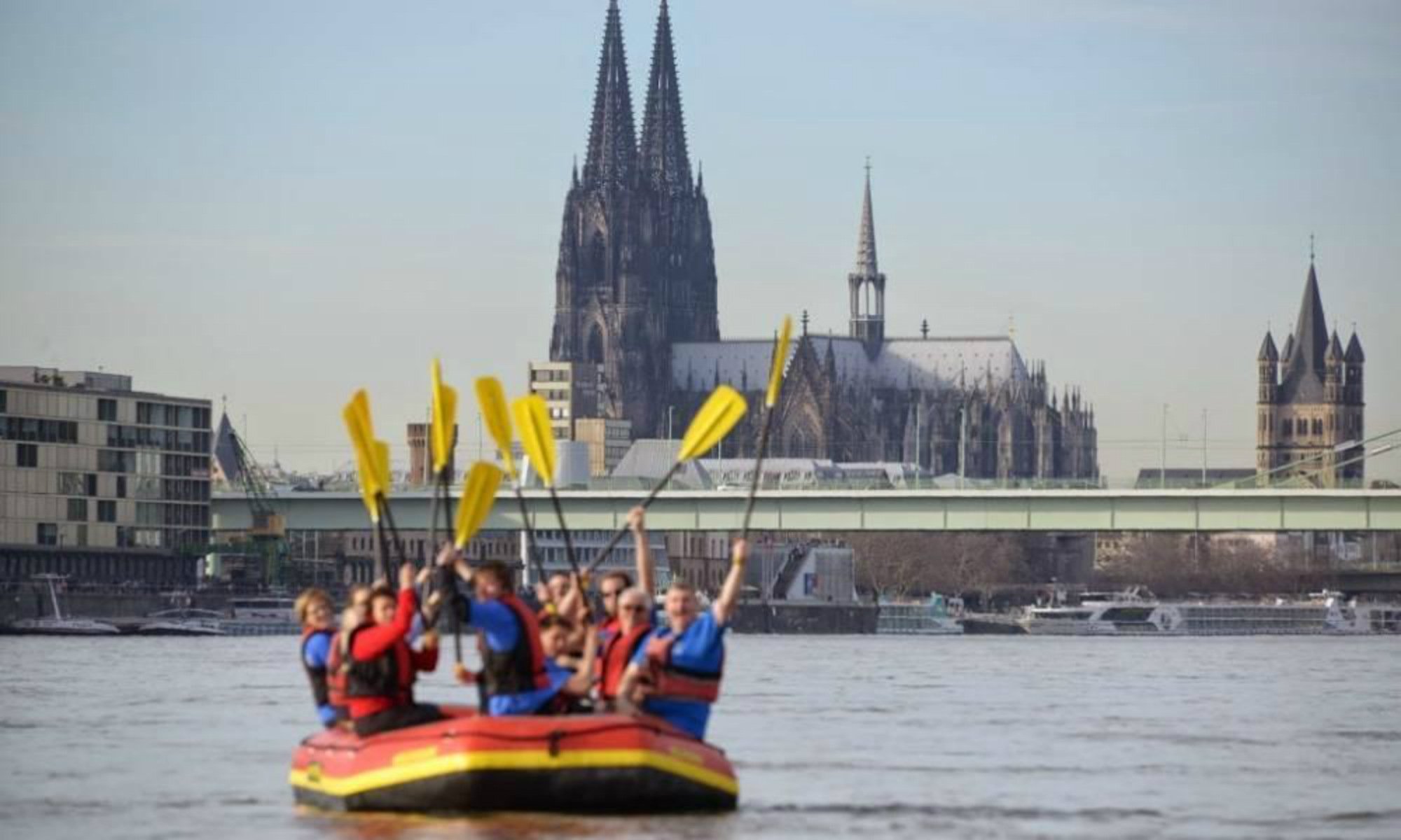 Eine Gruppe sitzt beim Rafting in NRW in einem Schlauchboot vor der Skyline von Köln.