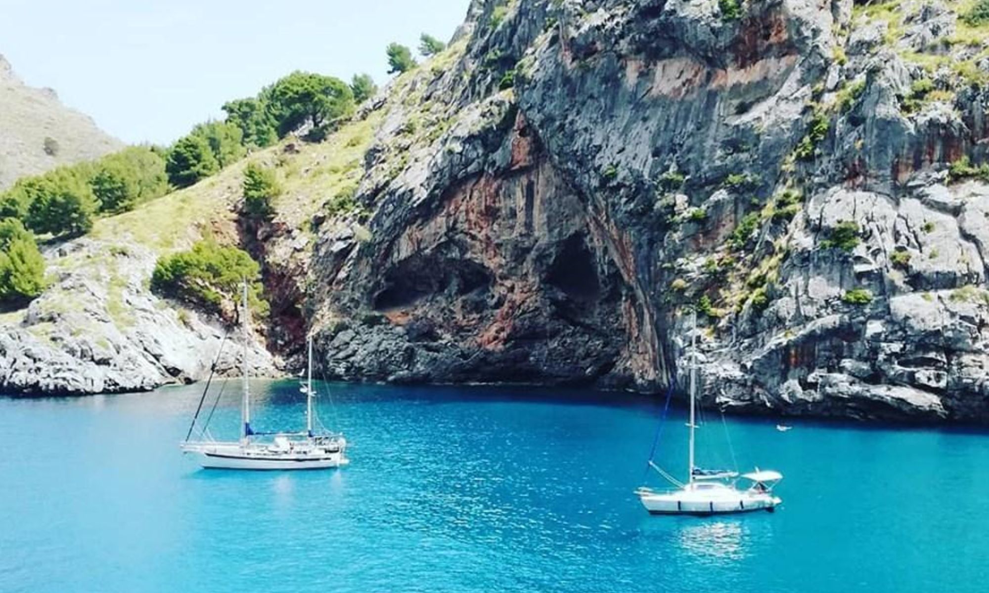 Zwei Segelboote ankern bei einer Bootstour in Mallorca vor einer zerklüfteten Felswand im türkisblauen Meer.