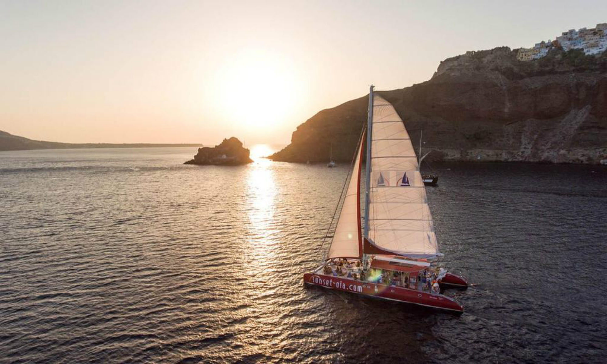 A catamaran sails around the island of Santorini at sunset.