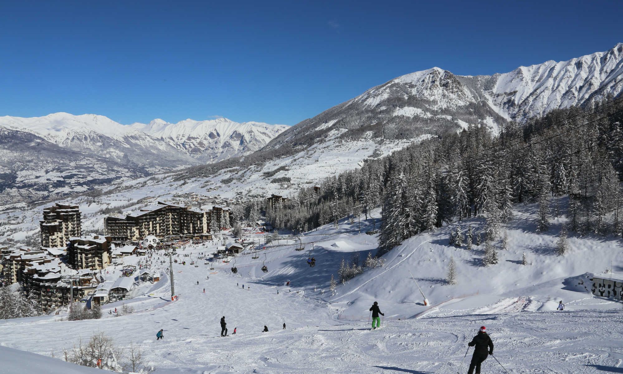 Des skieurs descendent une piste de ski en direction du village Les Orres 1650 sous un grand ciel bleu.