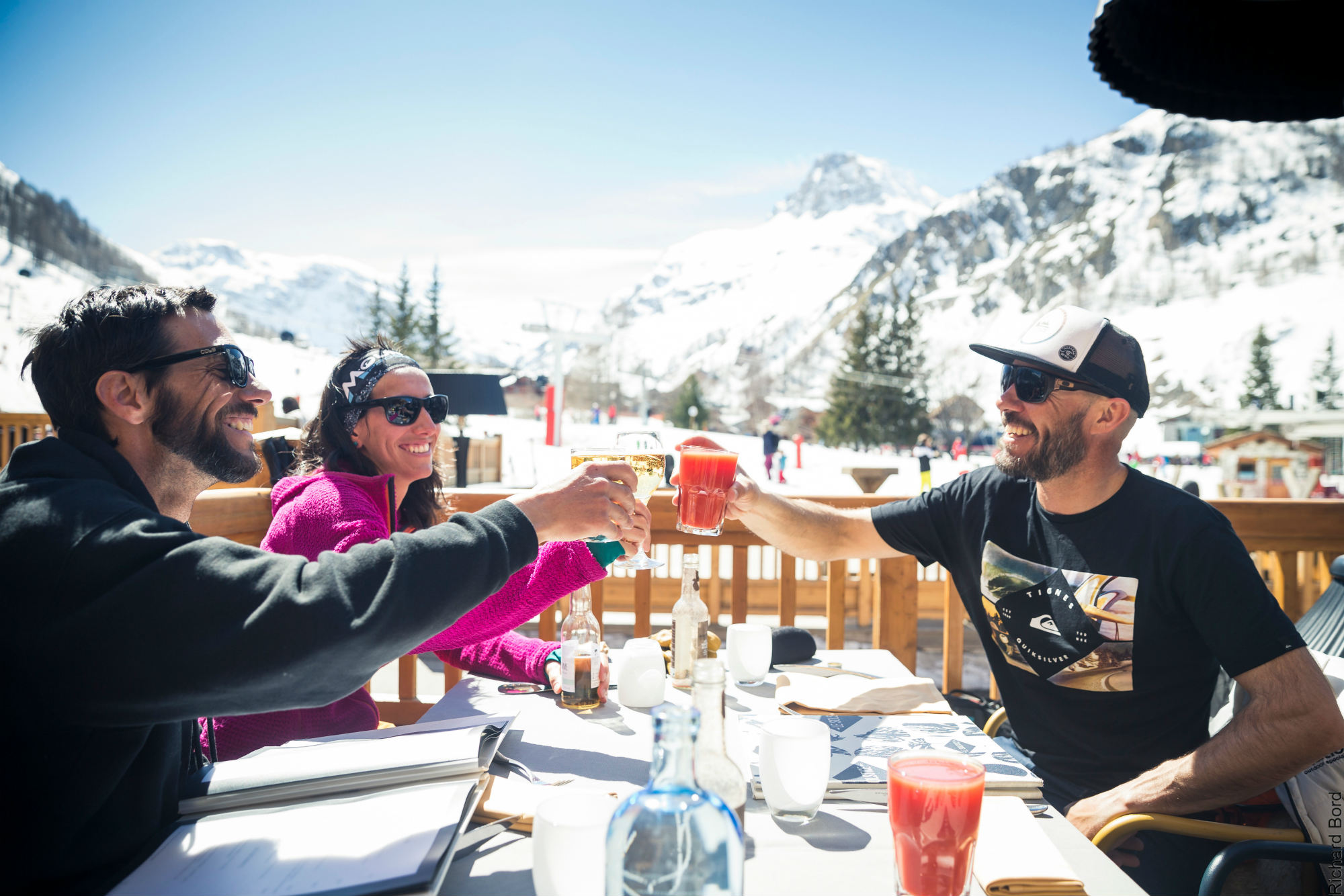 Trois amis trinquent à la terrasse ensoleillée d’un restaurant d’altitude à Val d’Isère.