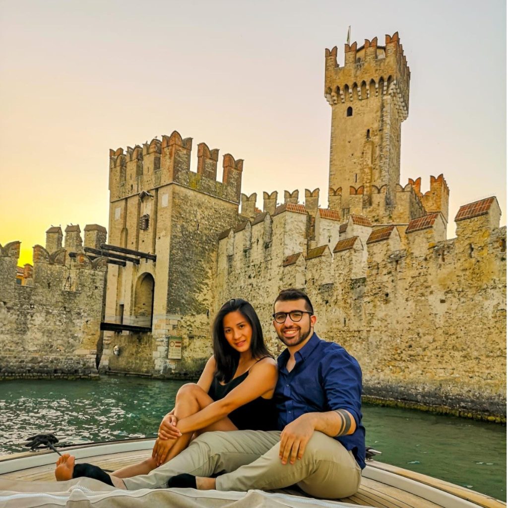 Una coppia posa su una barca al Lago di Garda al tramonto con il castello di Sirmione sullo sfondo.