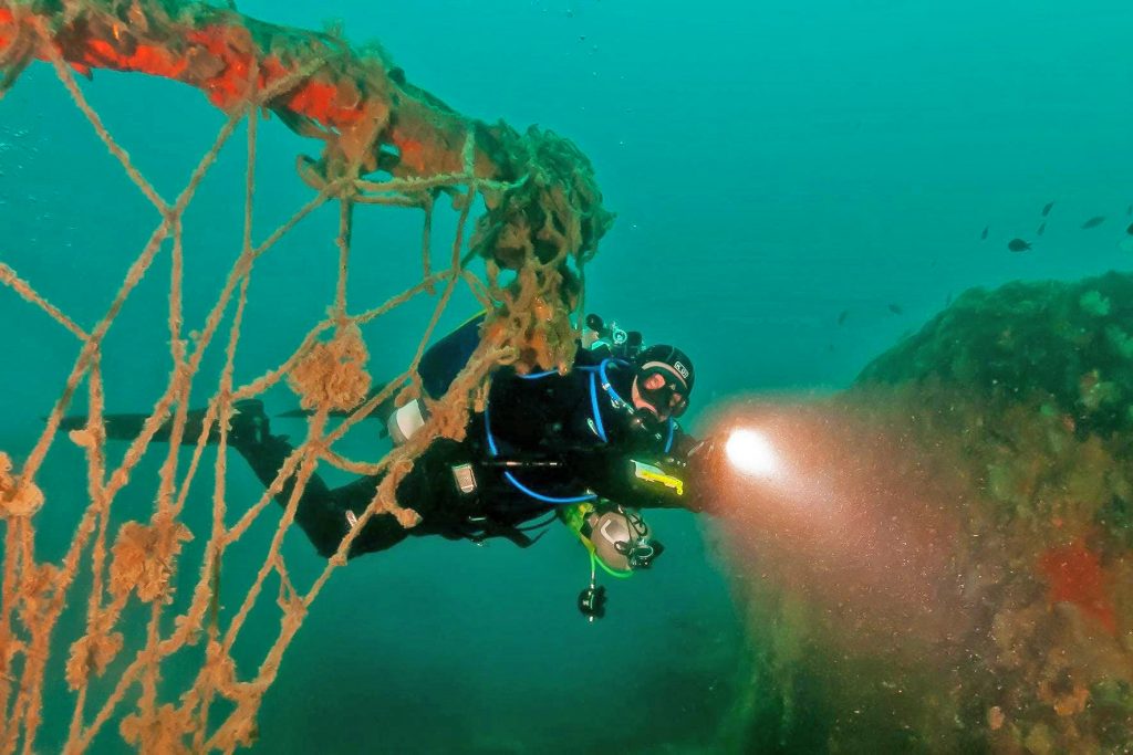 Un sommozzatore sta nuotando vicino a un relitto di nave a Pola, e illumina con la sua torcia le reti da pesca.