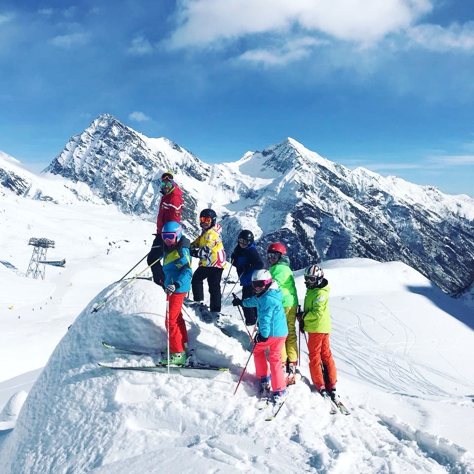 Bambini in piedi su un masso innevato con maestro di sci e montagne sullo sfondo.
