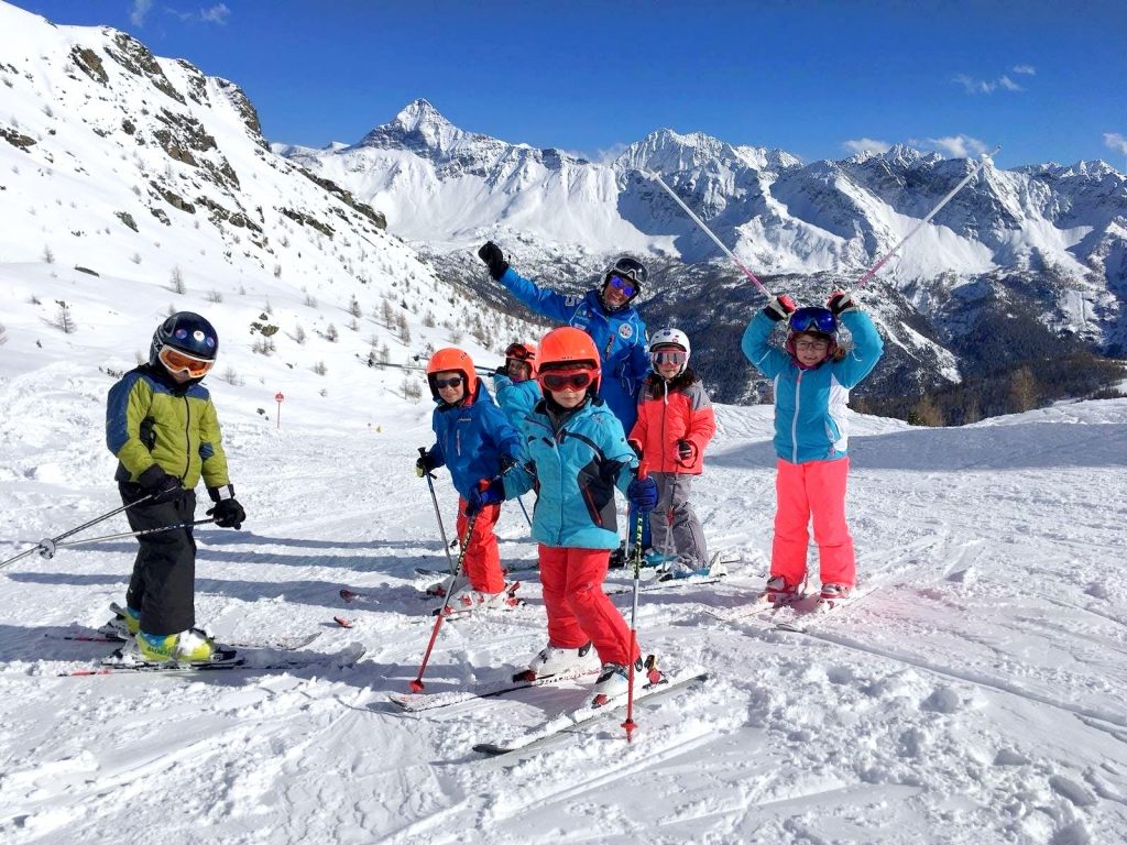Bambini in piedi sulla neve con maestro di sci e montagne sullo sfondo.
