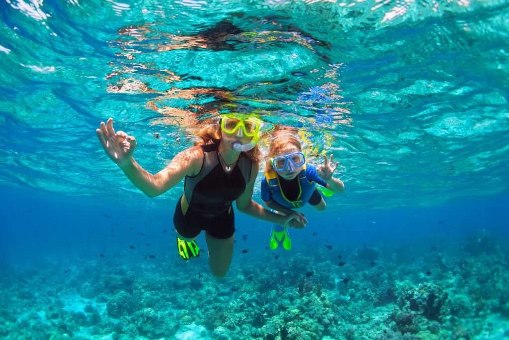 Mamma e figlia che fanno snorkeling nell'acqua blu e fanno ok con le mani alla fotocamera.