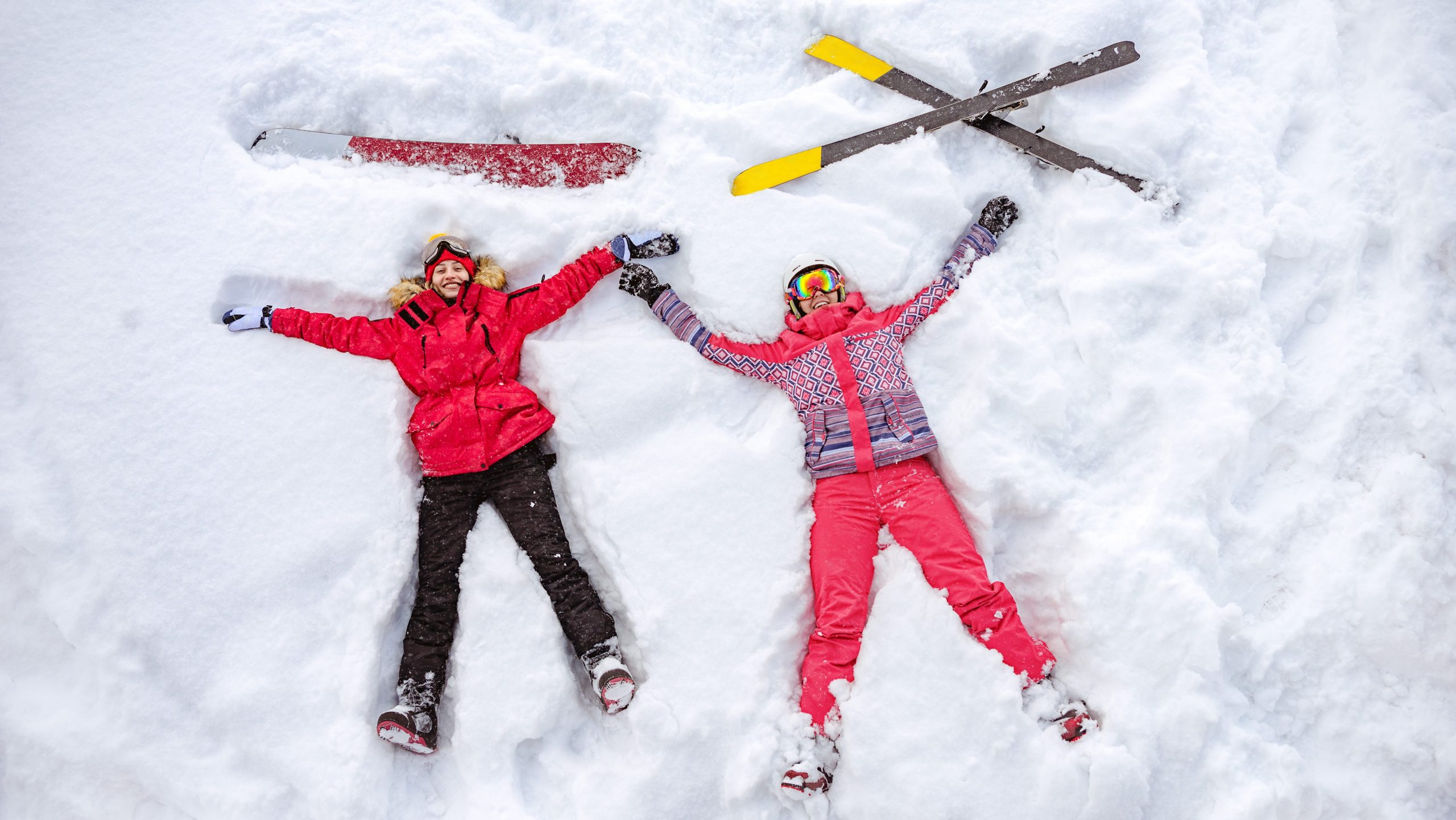 Eine Skifahrerin und eine Snowboarderin mit bunter Kleidung liegen im Schnee und lachen in die Kamera.