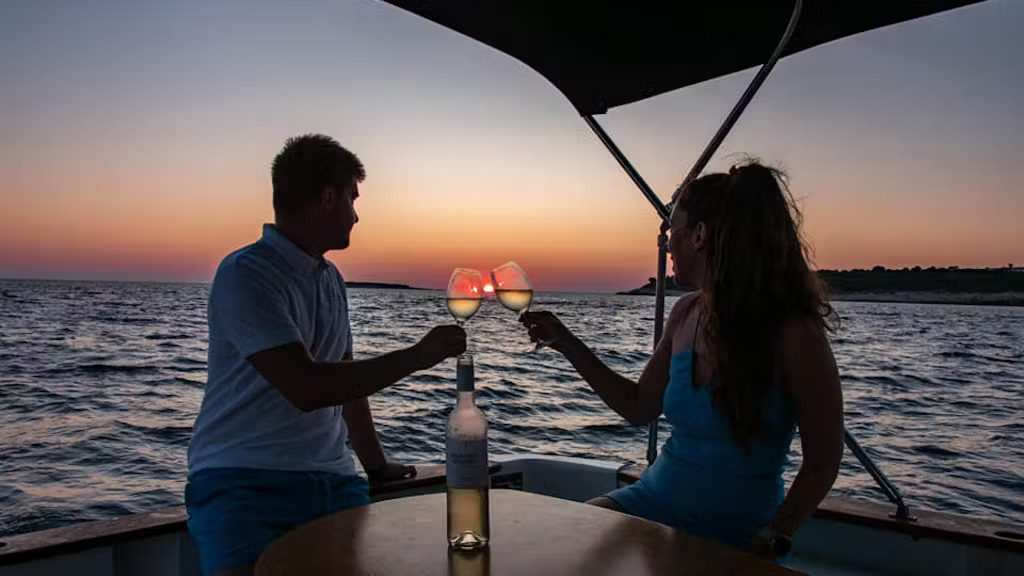 A couple clinks glasses while enjoying a romantic sunset on a boat, surrounded by calm waters and a colorful sky.